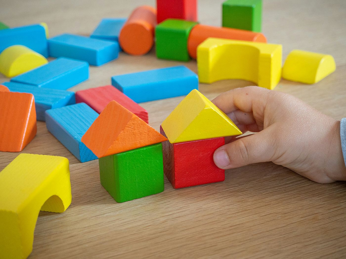 Eine Kinderhand setzt ein gelbes Dach auf ein kleines Haus aus bunten Holzbauklötzen